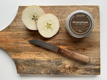 Lade das Bild in den Galerie-Viewer, Kramerhonig Bio-Holzpflege in der Dose legt zusammen mit einem angeschnittenen Apfel und einem Messer mit abgenutztem Holzgriff auf einem Holzbrett.
