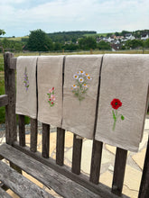 Lade das Bild in den Galerie-Viewer, Vier handbestickte Geschirrtücher aus 100 % Reinleinen, mit den Blumenmotiven Glockenblume, Heckenrose, Kamille und Mohn hängen nebeneinander über einer Holzbank vor natürlicher Landschaft.

