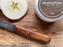 Lade das Bild in den Galerie-Viewer, Kramerhonig Bio-Holzpflege in der Dose legt zusammen mit einem angeschnittenen Apfel und einem mit der Holzpflge behandeltem Messer  mit samtglänzenden Holzgriff auf einem Holzbrett.

