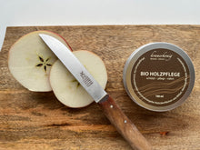 Lade das Bild in den Galerie-Viewer, Nahaufnahme Kramerhonig Bio-Holzpflege in der Dose zusammen mit einem angeschnittenen Apfel und einem Messer  mit abgenutztem Holzgriff auf einem Holzbrett.
